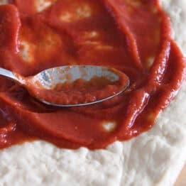 A metal spoon spreading pizza sauce on flattened, uncooked pizza dough.