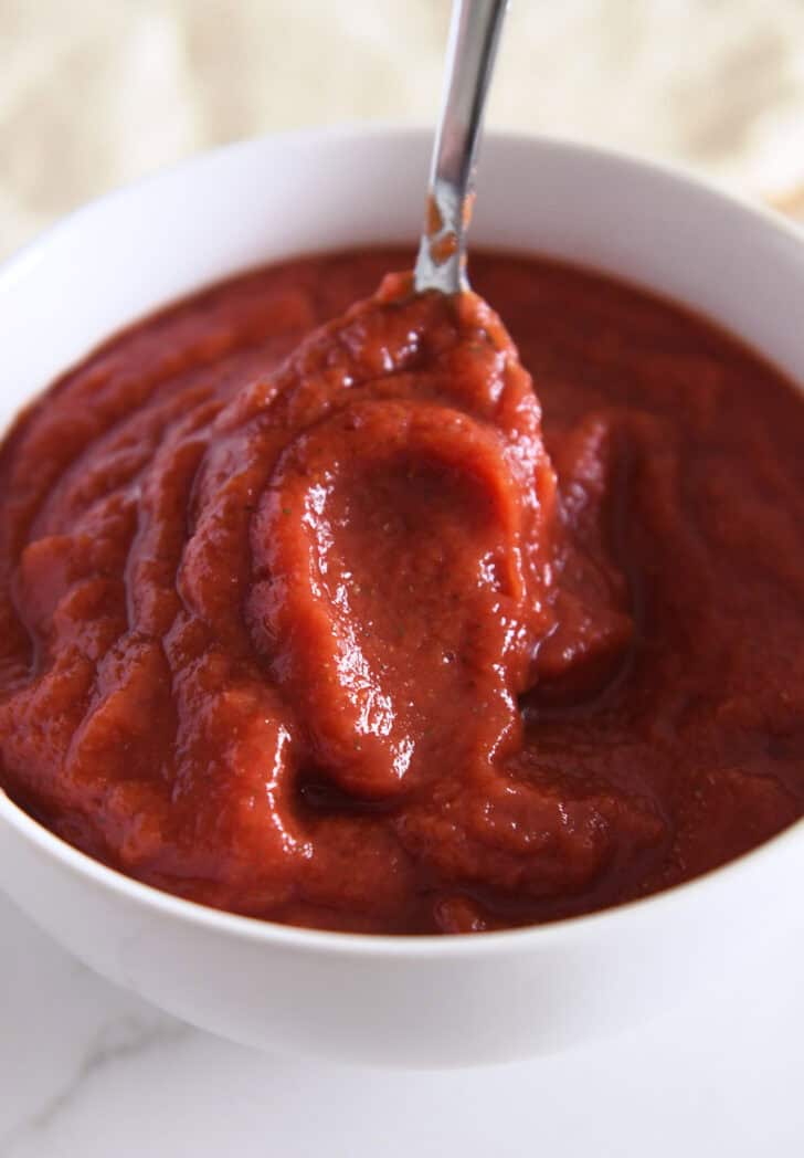 Metal spoon stirring pizza sauce in a white bowl.