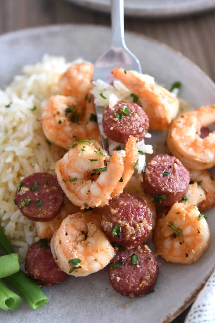 Fork scooping bite of shrimp and sausage on gray plate.