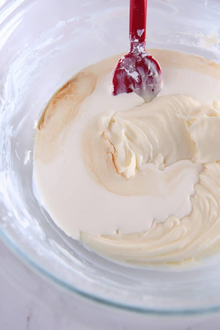 A glass bowl with a red spatula stirring a cream cheese mixture.