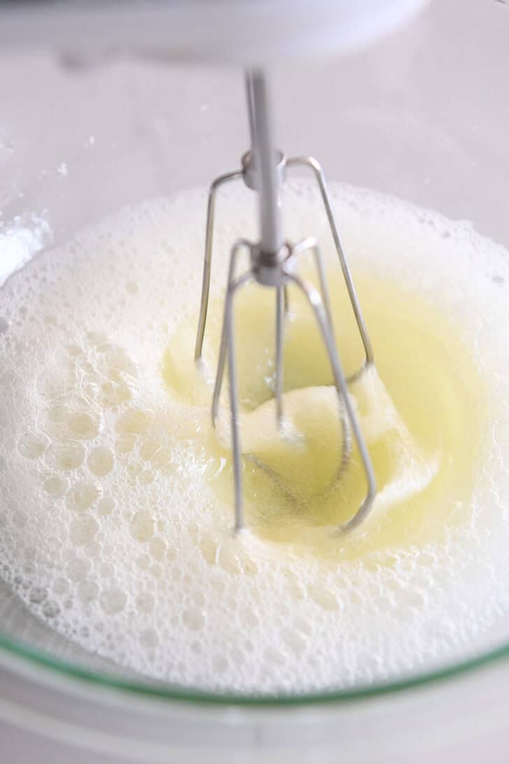 A frothy egg white mixture being whipped with a mixer in a glass bowl.