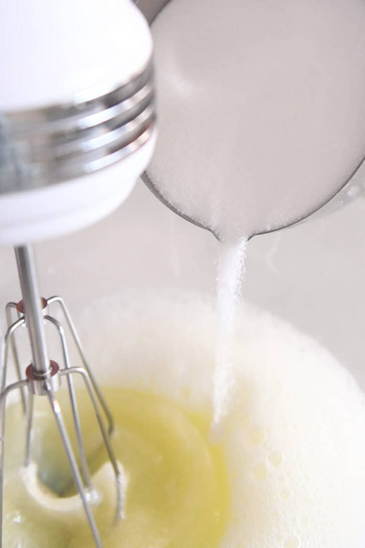 A metal measuring cup with sugar pouring from it into a glass bowl with egg whites that are being whipped.