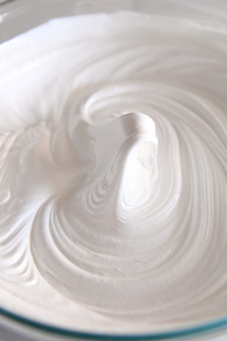 A glass bowl of egg whites with a stiff peak in the middle of the bowl.