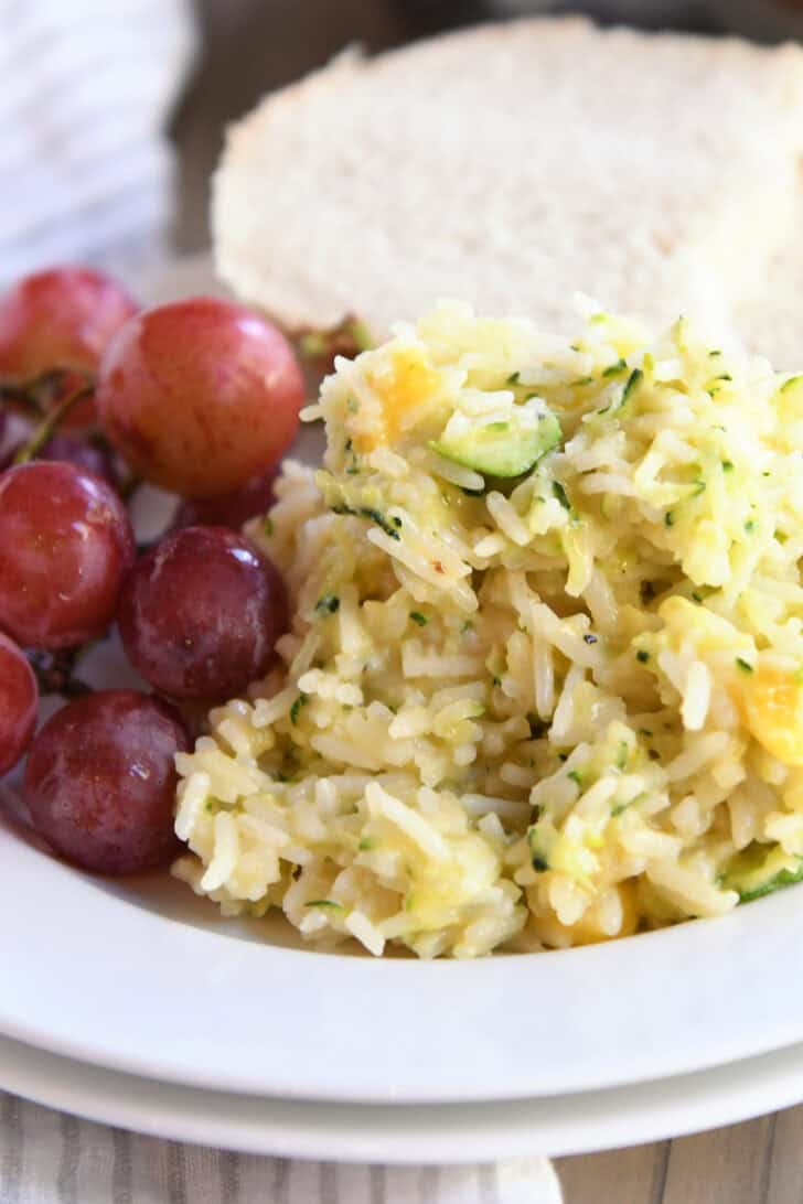 A white plate with cheesy zucchini rice, red grapes, and a slice of bread.