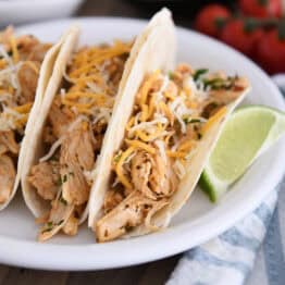 A white plate with three shredded chicken tacos topped with cheese in flour tortillas and a lime wedge next to the tacos.