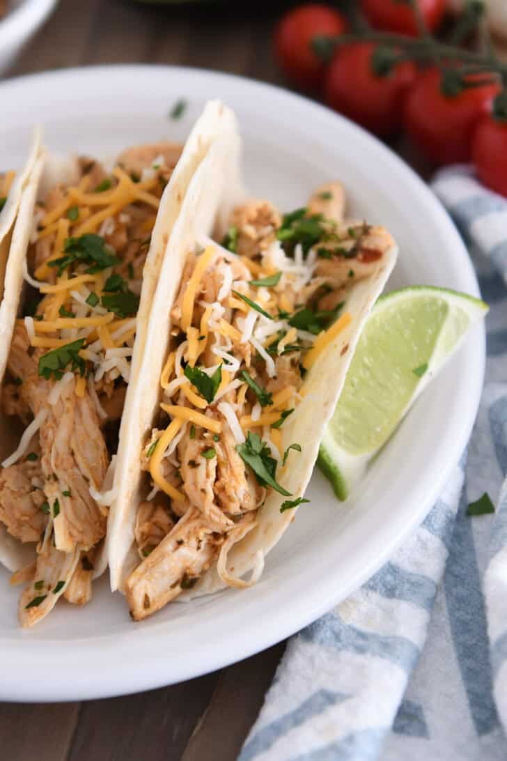 A white plate with assembled shredded chicken tacos topped with cheese and chopped cilantro.