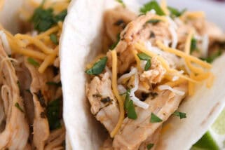 A white plate with two assembled shredded chicken tacos topped with cheese and chopped cilantro.