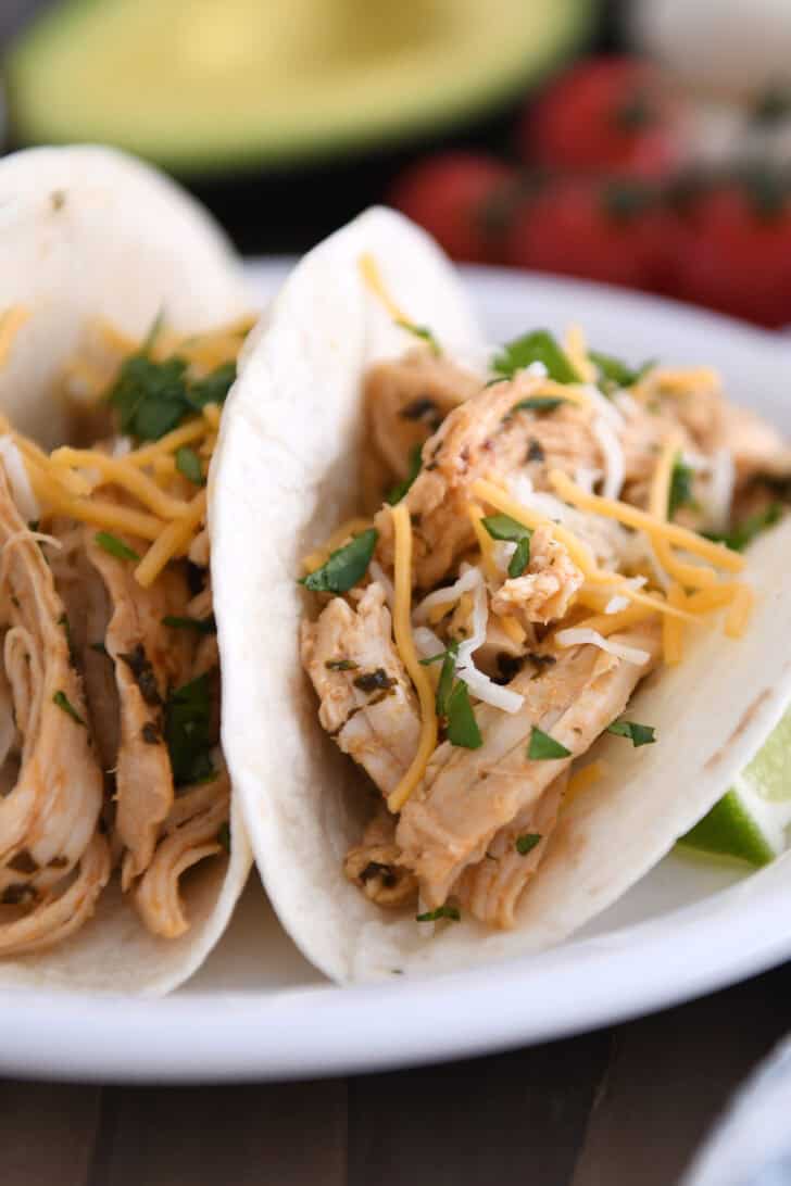 A white plate with two assembled shredded chicken tacos topped with cheese and chopped cilantro.