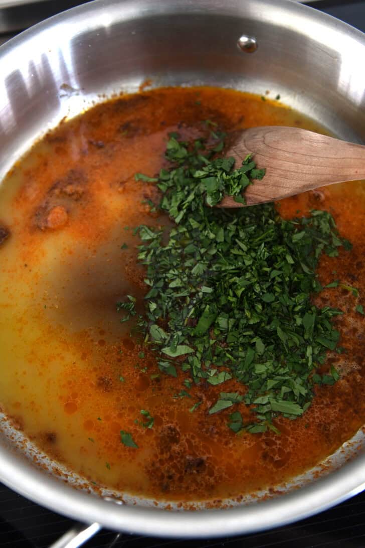 Top down view of a metal pot with an orange chipotle sauce, chopped cilantro, and a wooden spoon about to stir it.