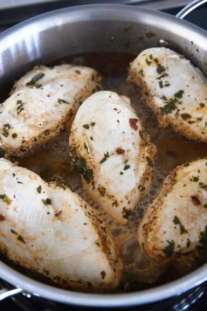 Top down view of five cooked chicken breasts simmering in a sauce on the stove.