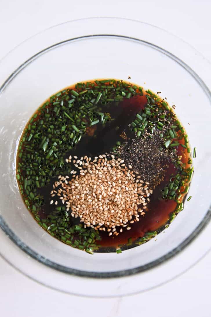 Top down view of glass bowl with soy sauce, sesame seeds, pepper and green onions.