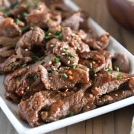 White tray with cooked pieces of thinly sliced beef, green onions and sesame seeds.