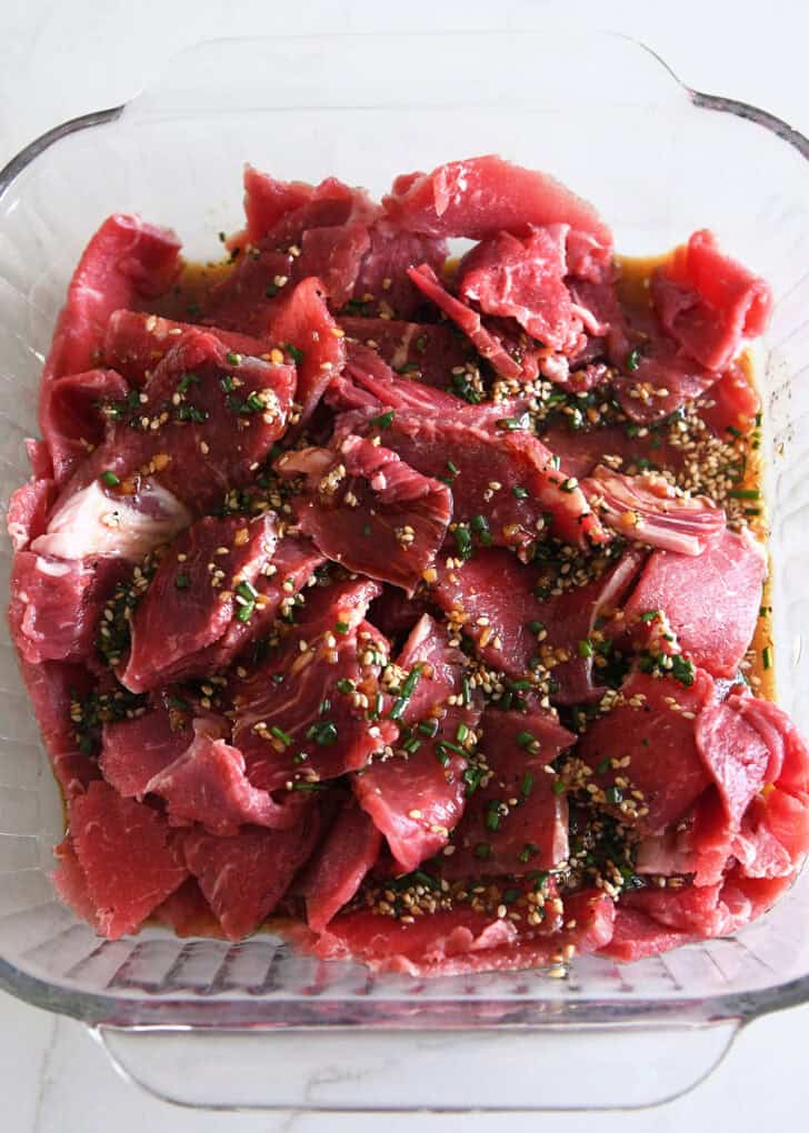 Top down view of square baking dish with thinly sliced beef and soy sauce, green onions and sesame seeds.