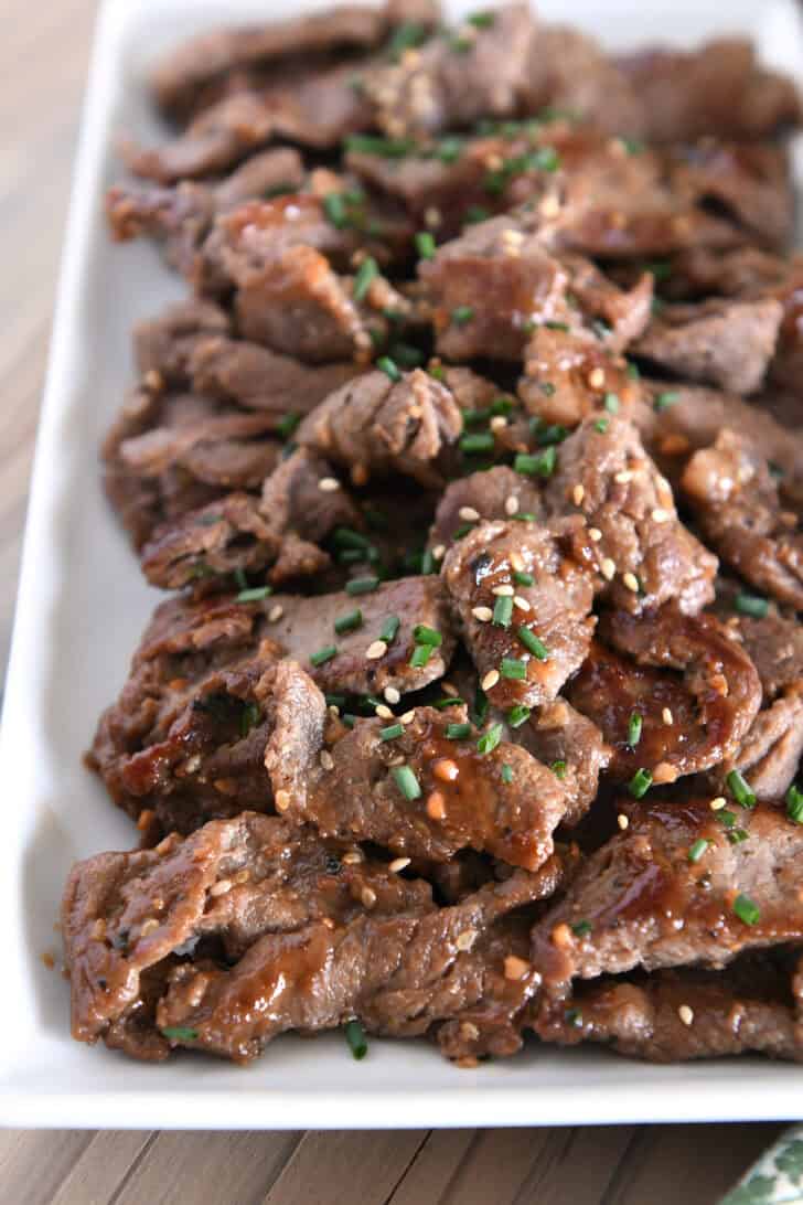 White tray piled with cooked pieces of thin beef, green onions, sauce and sesame seeds.