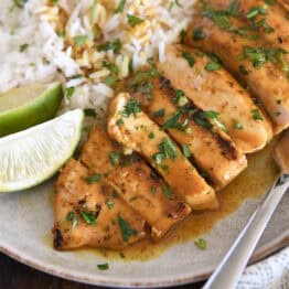 Grilled chicken breast slices with sauce, rice and fresh limes on gray plate.