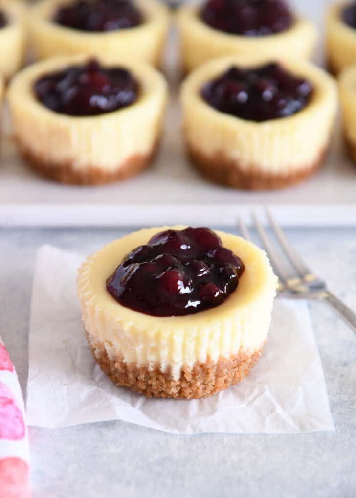 One mini cheesecake with huckleberry topping with tray of mini cheesecakes in background.
