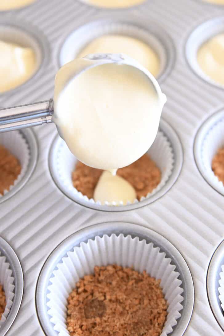 Scooping cheesecake batter into muffin liners with graham cracker crust.
