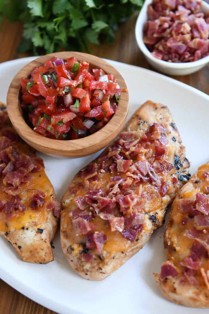 Top down view of grilled chicken breasts topped with melted cheese and bacon and a wooden bowl filled with pico de gallo on a white plate with the chicken.