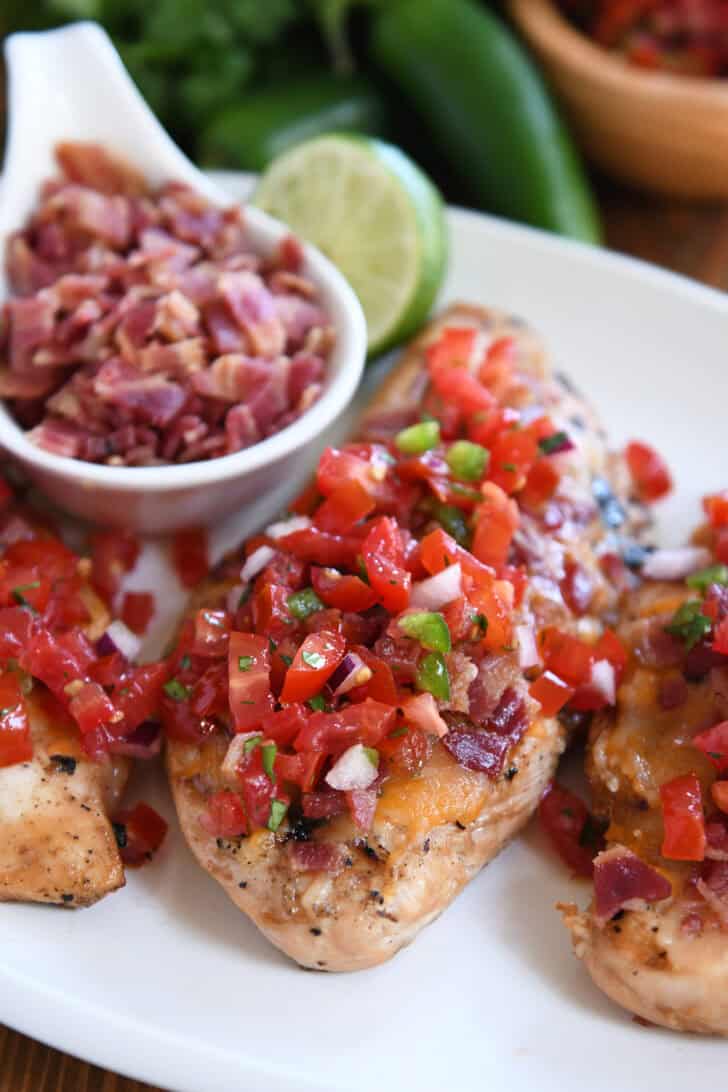 Grilled chicken breasts topped with pico de gallo on a white plate with a bowl of chopped bacon and a half of a lime.