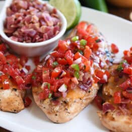 Grilled chicken breasts topped with pico de gallo on a white plate with a bowl of chopped bacon and a half of a lime in the background.
