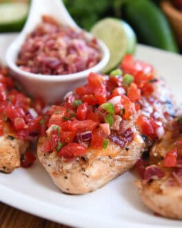 Grilled chicken breasts topped with pico de gallo on a white plate with a bowl of chopped bacon and a half of a lime behind the chicken.