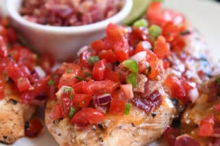 Grilled chicken breasts topped with pico de gallo on a white plate with a bowl of chopped bacon and a half of a lime behind the chicken.