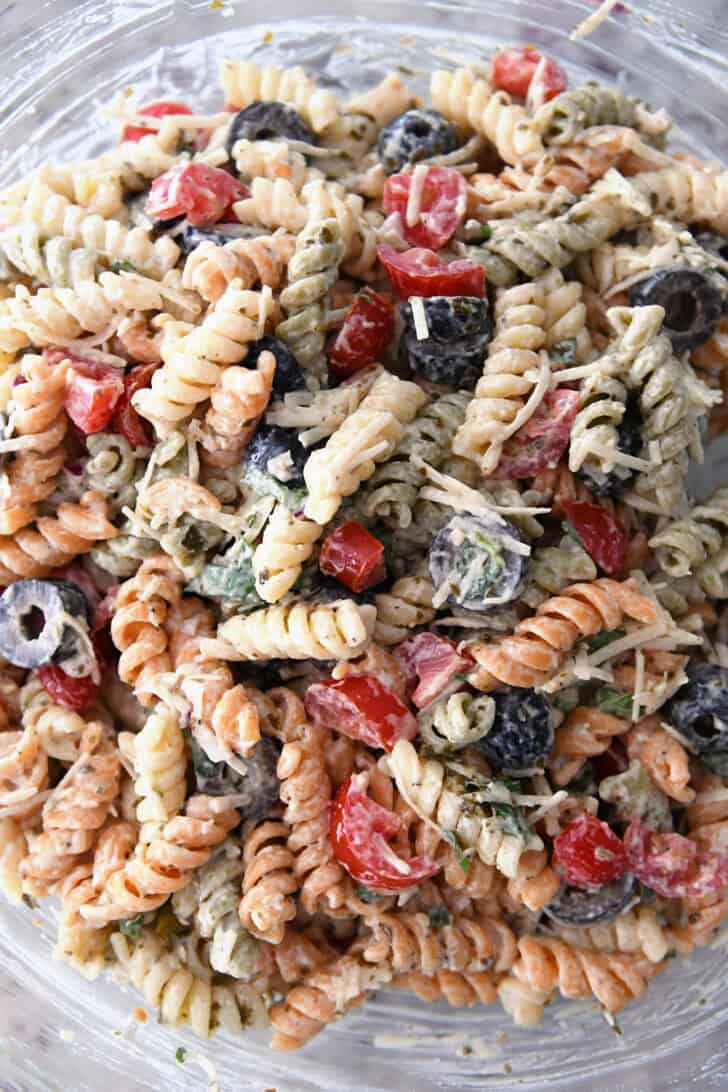Top down view of tri-color rotini pasta in bowl with salad dressing, olives, tomatoes and parmesan cheese.