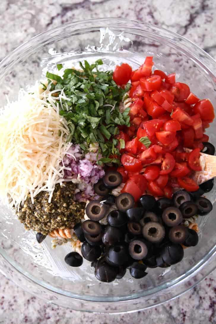 Top down view of olives, pesto, red onions, tomatoes, basil and parmesan cheese in glass bowl.