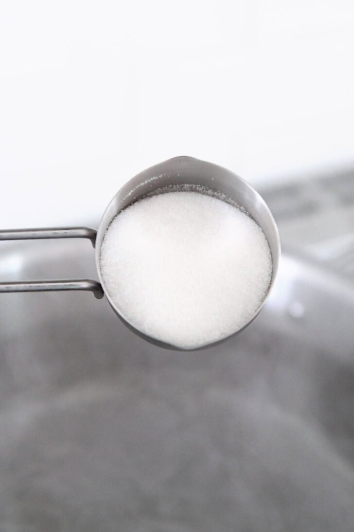 A metal measuring cup with sugar over a metal pot with boiling water.