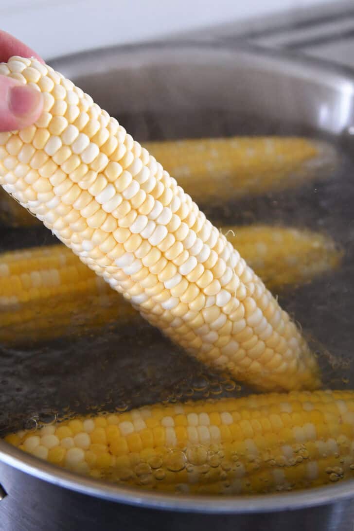 A hand putting an ear of corn into a pot with other boiling ears of corn.