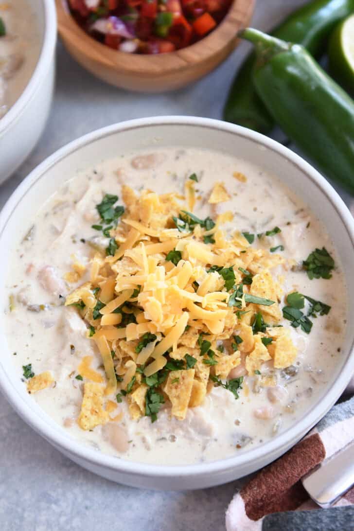 Top down view of a white bowl of white chicken chili topped with shredded cheddar cheese, chopped cilantro, and crushed tortilla chips.