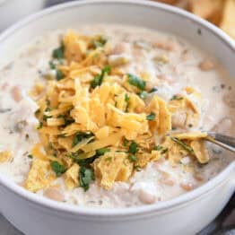 A white bowl of white chicken chili topped with shredded cheddar cheese, chopped cilantro, and crushed tortilla chips.