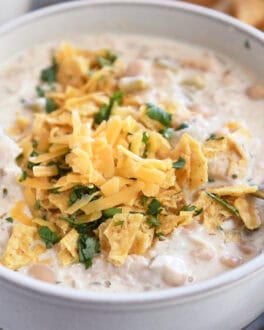 A white bowl of white chicken chili topped with shredded cheddar cheese, chopped cilantro, and crushed tortilla chips.
