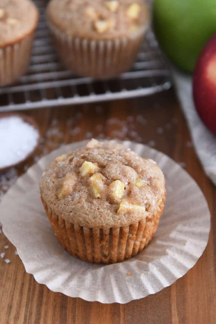 Unwrapped apple muffin on paper liner with coarse sugar sprinkled behind and apples in the background.
