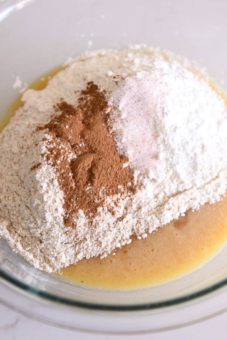 Flour, whole wheat flour, cinnamon and salt on top of eggs and oil in glass bowl.