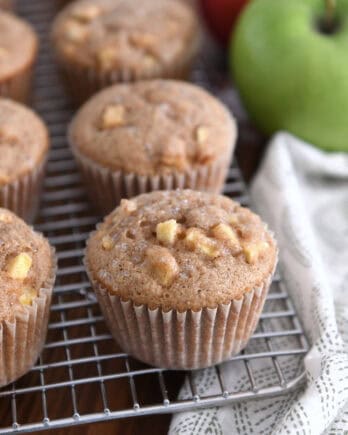 Several apple cinnamon muffins on cooling rack with apples in the background.