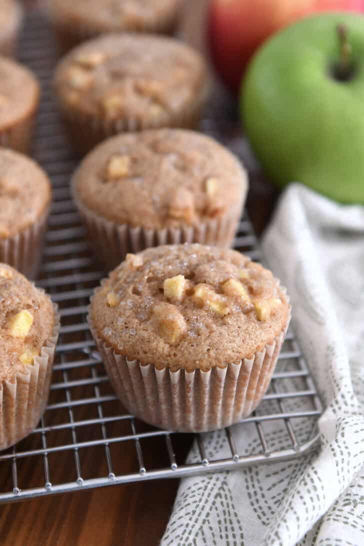 Several apple cinnamon muffins on cooling rack with apples in the background.