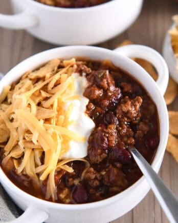 Spoon scooping bite of beef and bean tomato chili in sour cream, shredded cheese and corn chips.
