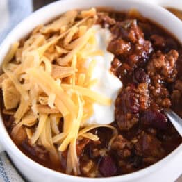 White bowl with handles and spoon scooping bite of beef and bean chili with shredded cheese, sour cream and corn chips.