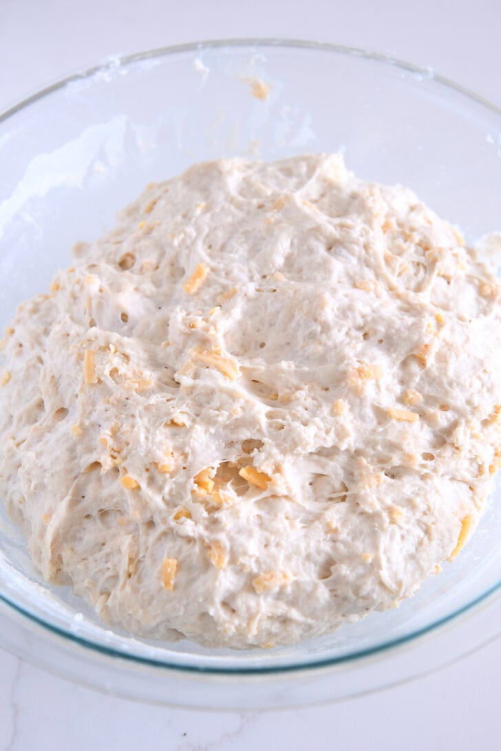 Mixed and risen cheesy peasant bread dough in glass bowl.