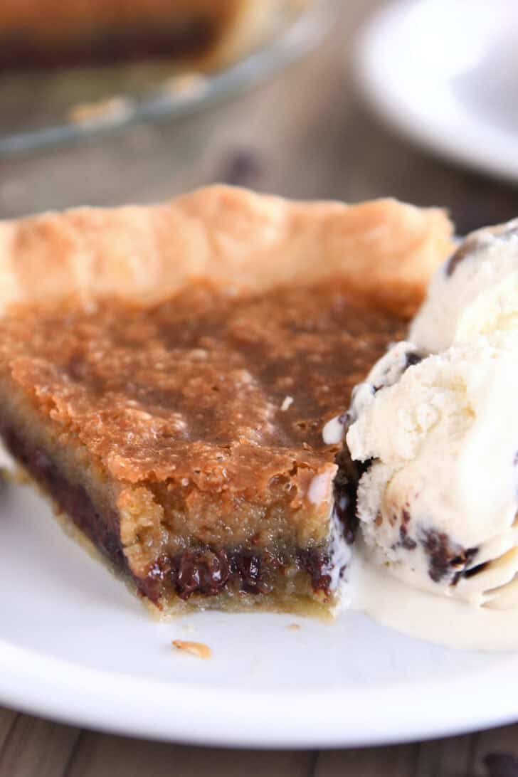 A slice of chocolate chip cookie pie with a bite taken out on a white plate. A scoop of ice cream is next to the pie.