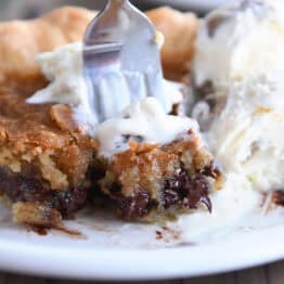 A white plate with a slice of chocolate chip cookie pie and a fork taking a bite out of the slice.