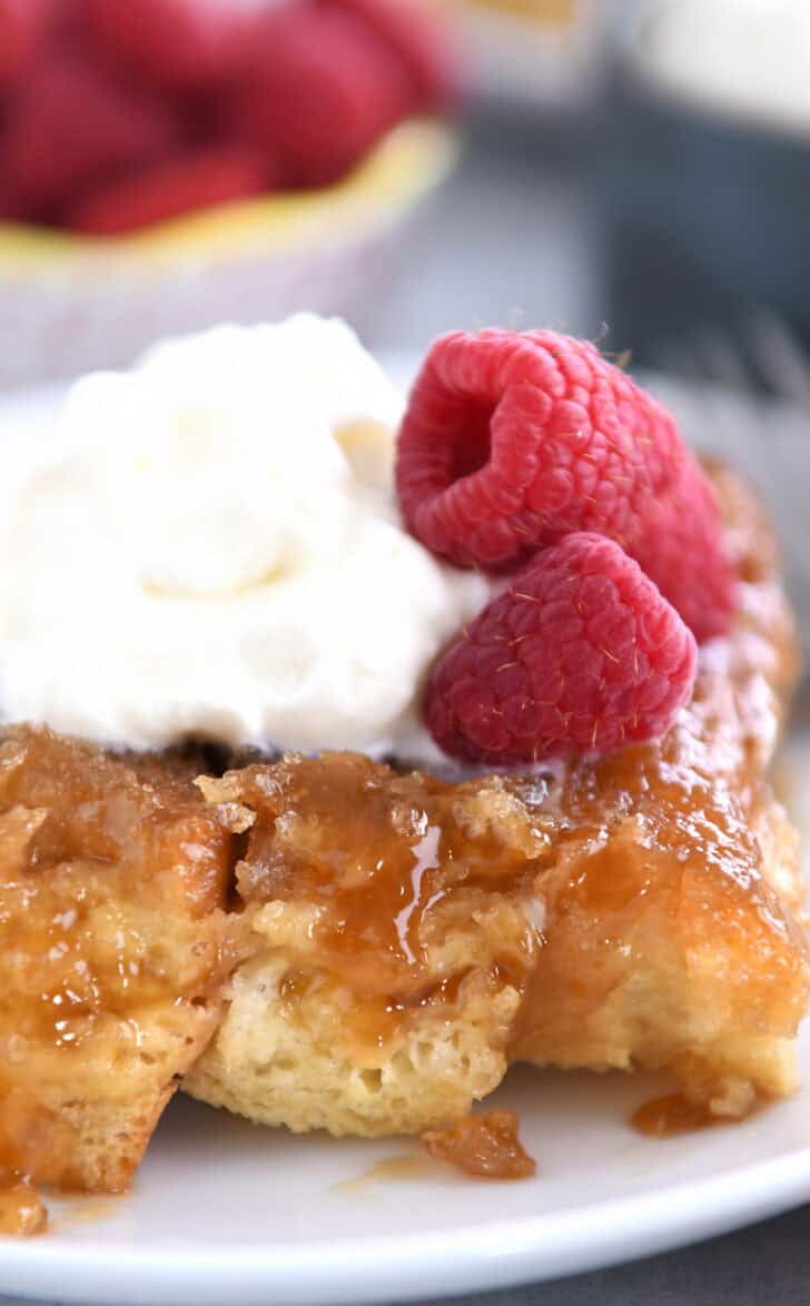 A white plate with a piece of french toast bake topped with whipped cream and fresh raspberries.