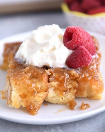 A white plate with a piece of french toast bake topped with whipped cream and fresh raspberries.