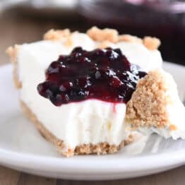 A slice of cheesecake with a dollop of blueberry sauce on top, on a white plate. A fork has a bite of the cheesecake on it next to the slice.