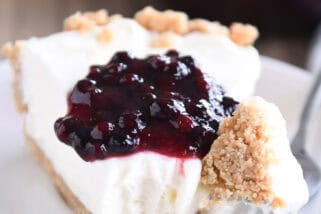 A slice of cheesecake with a dollop of blueberry sauce on top, on a white plate. A fork has a bite of the cheesecake on it next to the slice.