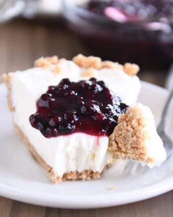 A slice of cheesecake with a dollop of blueberry sauce on top, on a white plate. A fork has a bite of the cheesecake on it next to the slice.