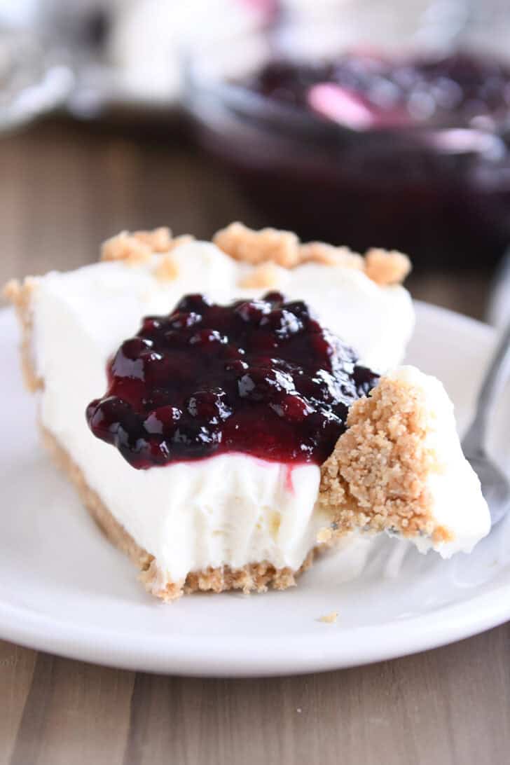 A slice of cheesecake with a dollop of blueberry sauce on top, on a white plate. A fork has a bite of the cheesecake on it next to the slice.