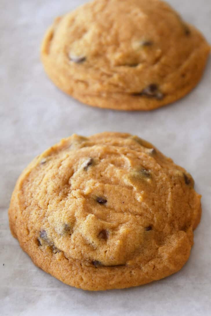 Top down view of two pumpkin chocolate chip cookies spaced a few inches apart on a piece of parchment paper.