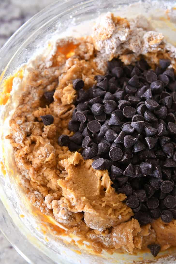 Top down view of a glass bowl full of pumpkin cookie batter with a heaping scoop of chocolate chips on top ready to be mixed in.